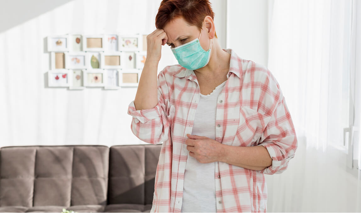 Woman wearing a surgical mask holding her abdomen and head, depicting common symptoms of colorectal cancer such as abdominal pain, nausea, and fatigue